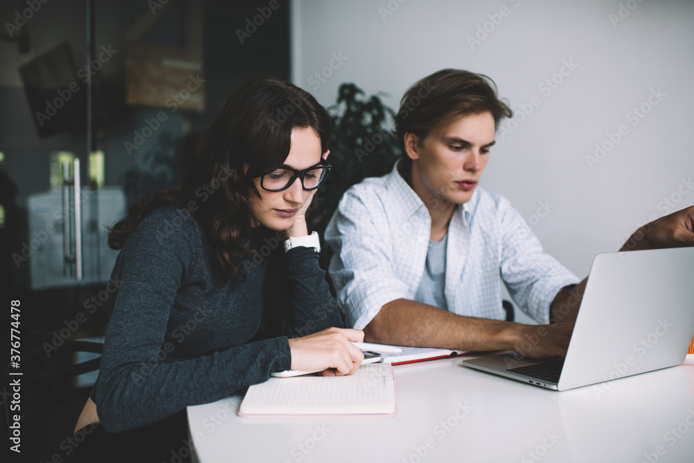 Serious clever woman in eyewear writing in notepad doing homework with caucasian man, pensive colleagues cooperating on business planning using laptop computer for researching in coworking office. Serious clever woman in eyewear writing in notepad doing homework with caucasian man, pensive colleagues cooperating on business planning using laptop computer for researching in coworking office.