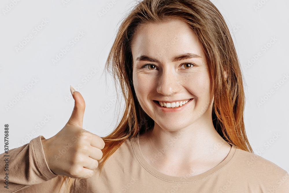 Beautiful young girl smiling and shows thumbs up gesture on a white isolated background. A woman points to an idea, a place for advertising. Positive brunette in a beige jumper. Beautiful young girl smiling and shows thumbs up gesture on a white isolated background. A woman points to an idea, a place for advertising. Positive brunette in a beige jumper.