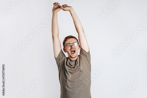 Good morning. The sleepy man yawns, pulls his arms up and stretches. Boredom and sleepiness. Young handsome man with glasses a brunette beard posing on gray background. Place for advertising.