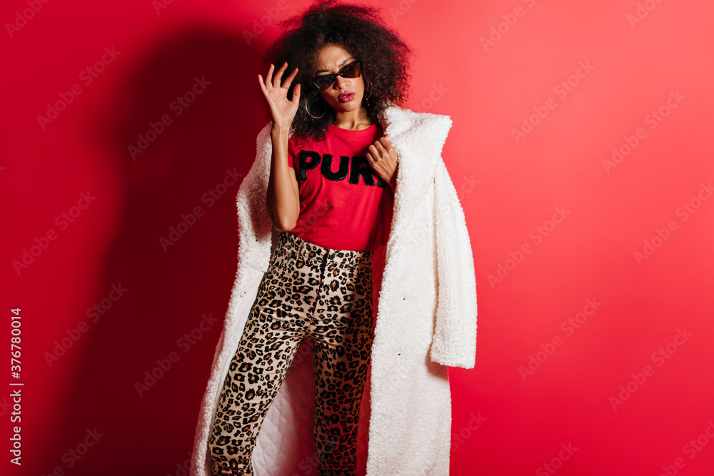 Sensual stylish girl in sunglasses posing on red background. Slim trendy african lady in coat chilling in studio. Sensual stylish girl in sunglasses posing on red background. Slim trendy african lady in coat chilling in studio.
