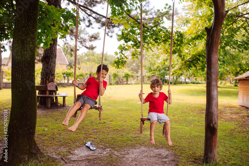 Two cute caucasian brother boys in red T-shirts are swinging on a swing in the park