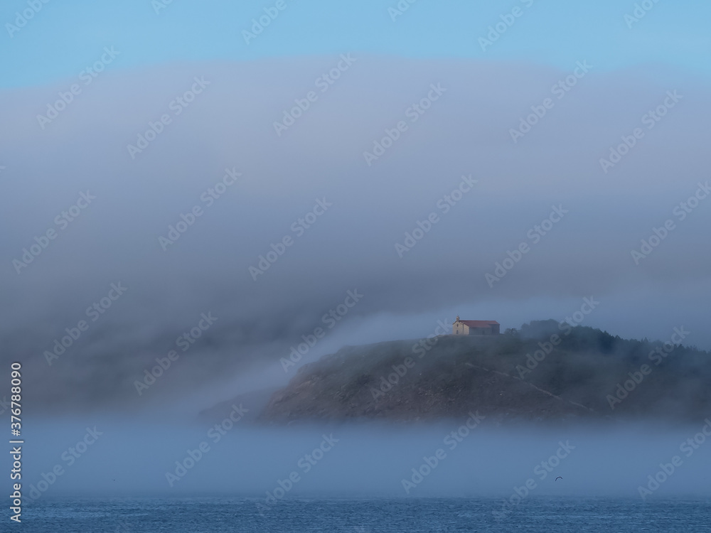 Landscape among low clouds where you can see a church on a mountain and the sea