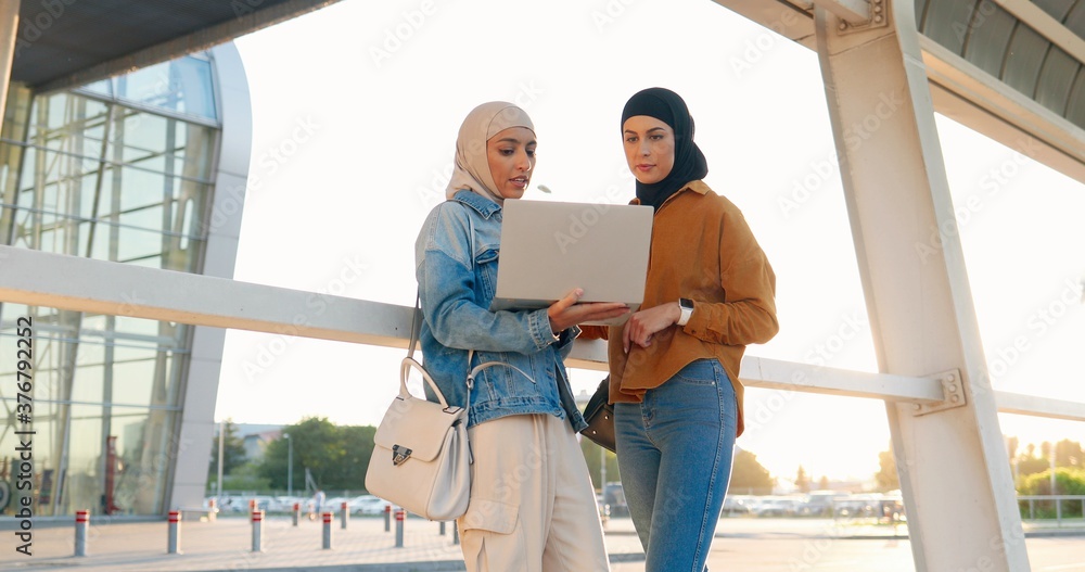 Two Caucasian young females friends in traditional Arabic headscarves standing at street and using laptop computer. Muslims friendly beautiful women alking and discussing something on screen.