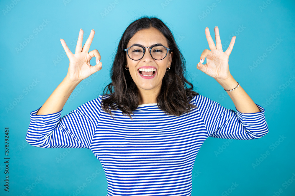 Pretty woman wearing glasses standing over insolated blue background doing okay sign with fingers and smiling, excellent symbol. Pretty woman wearing glasses standing over insolated blue background doing okay sign with fingers and smiling, excellent symbol.
