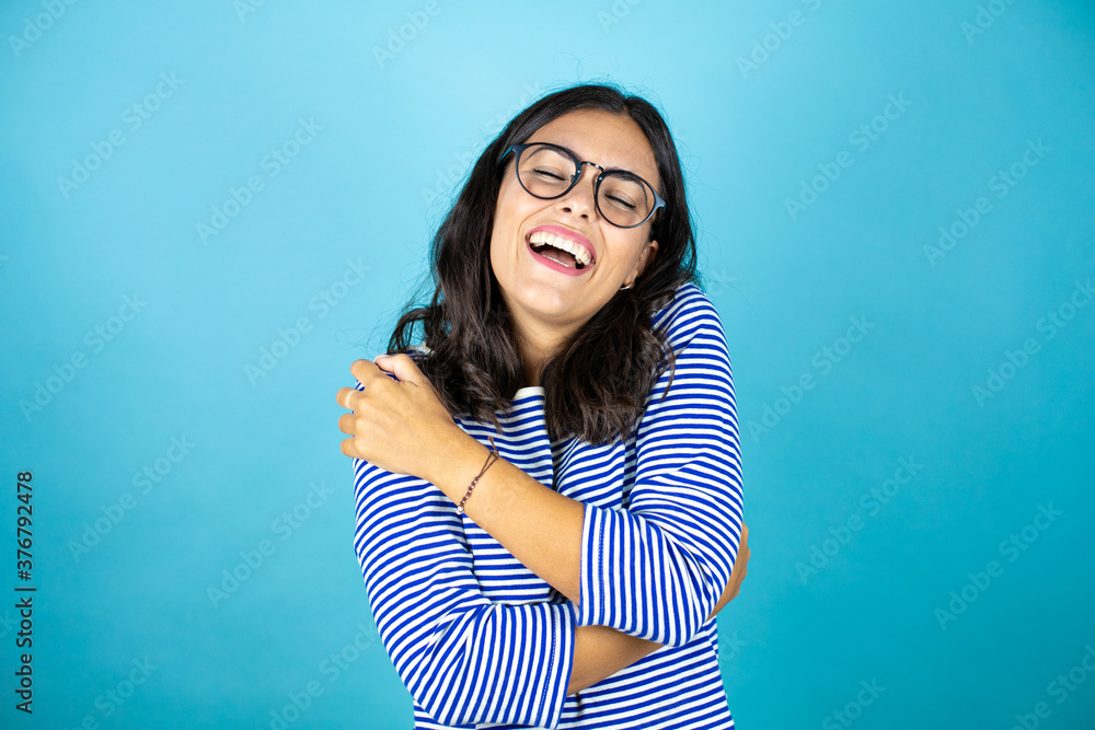 Pretty woman wearing glasses standing over insolated blue background hugging oneself happy and positive, smiling confident.