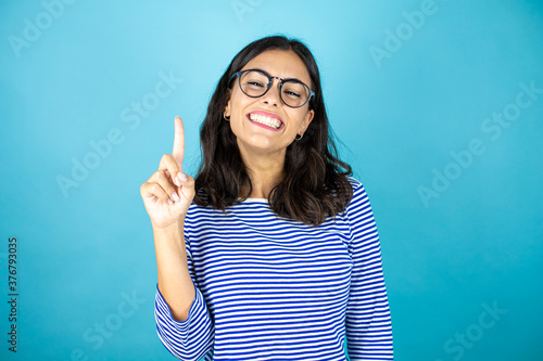 Pretty woman wearing glasses standing over insolated blue background showing and pointing up with fingers number one while smiling confident and happy.