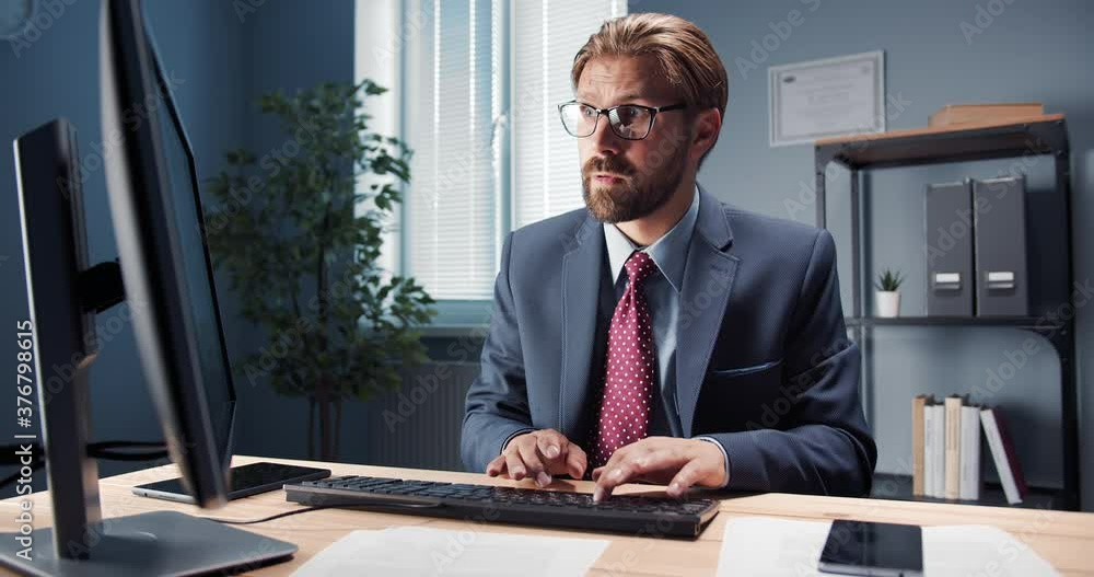 Handsome man wearing eyeglasses and business suit, sitting at table and typing on computer. Bearded serious person working at office with modern gadget. Handsome man wearing eyeglasses and business suit, sitting at table and typing on computer. Bearded serious person working at office with modern gadget.
