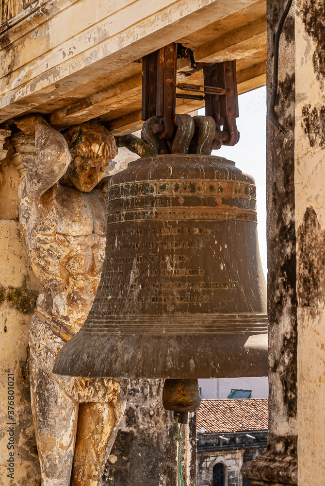 Obraz premium Leon, Nicaragua - November 27, 2008: Cathedral of Assumption, Asuncion.. Closeup of heavy dark metal old bell hanging in part of yellow stone towers, flanked by statue of man supporting beam. 