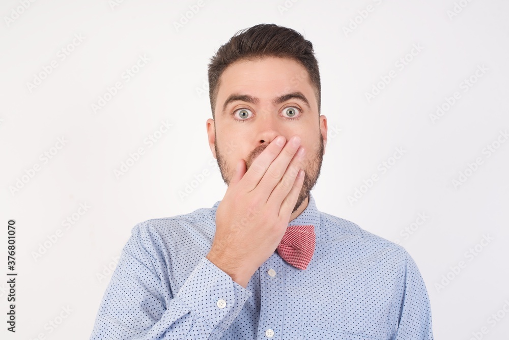 Emotional European man dressed in formal shirt and bow tie poses against white studio background gasps from astonishment, covers opened mouth with palm, looks shocked at camera. Reaction concept. Emotional European man dressed in formal shirt and bow tie poses against white studio background gasps from astonishment, covers opened mouth with palm, looks shocked at camera. Reaction concept.