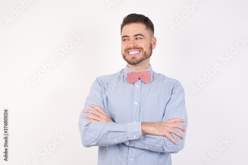 Dreamy rest relaxed European man dressed in formal shirt and bow tie poses against white studio background crossing arms, looks good