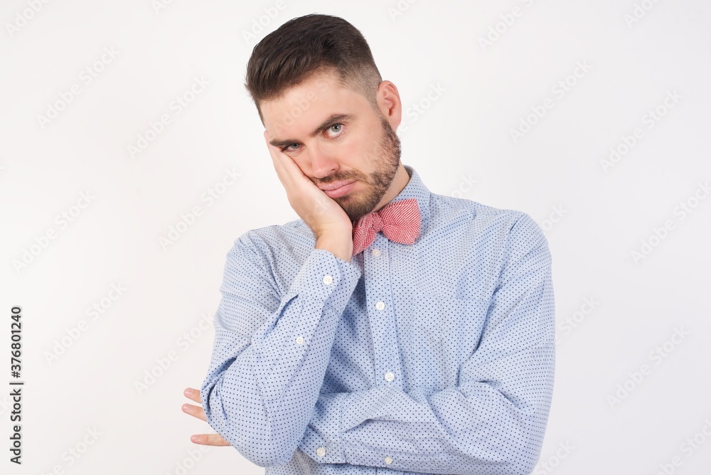 Very bored European man dressed in formal shirt and bow tie poses against white studio background holding hand on cheek while support it with another crossed hand, looking tired and sick, Very bored European man dressed in formal shirt and bow tie poses against white studio background holding hand on cheek while support it with another crossed hand, looking tired and sick,