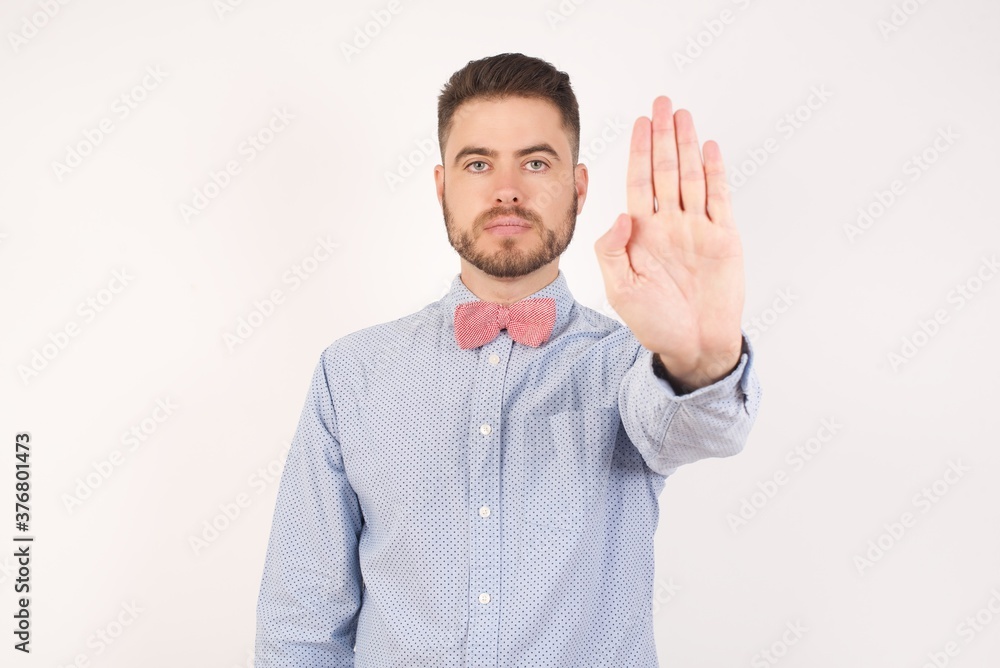 Fototapeta premium European man dressed in formal shirt and bow tie poses against white studio background doing stop gesture with palm of the hand. Warning expression with negative and serious gesture on the face