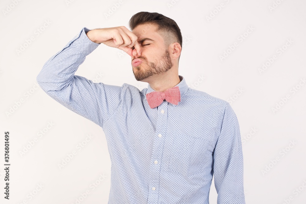 European man dressed in formal shirt and bow tie poses against white studio background smelling something stinky and disgusting, intolerable smell, holding breath with fingers on nose. Bad smell European man dressed in formal shirt and bow tie poses against white studio background smelling something stinky and disgusting, intolerable smell, holding breath with fingers on nose. Bad smell