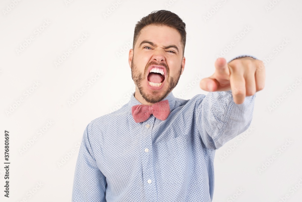 European man dressed in formal shirt and bow tie poses against white background pointing displeased and frustrated to the camera, angry and furious ready to fight with you. European man dressed in formal shirt and bow tie poses against white background pointing displeased and frustrated to the camera, angry and furious ready to fight with you.
