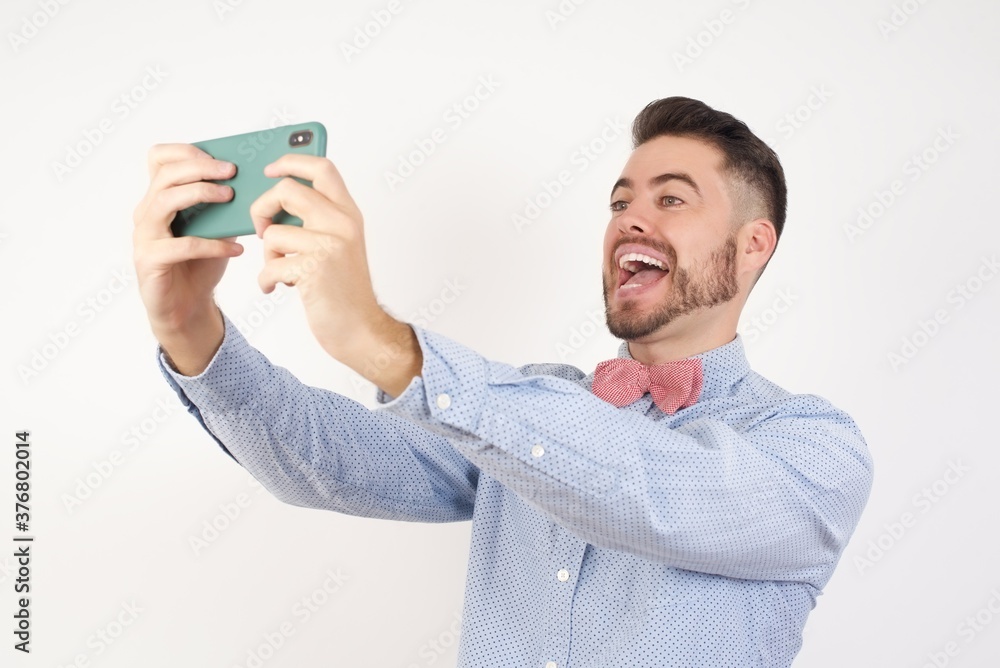 Close-up portrait European man dressed in formal shirt and bow tie poses against white studio backgroundtaking a selfie to post it on social media or having a video call with friends. Close-up portrait European man dressed in formal shirt and bow tie poses against white studio backgroundtaking a selfie to post it on social media or having a video call with friends.