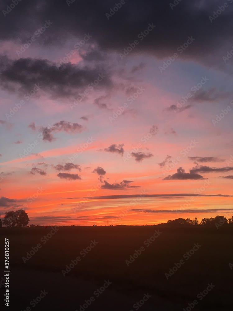 Brilliant sunset against stark country landscape that glows in yellows, oranges, peaches, light blue against pink, violet, gray and black clouds. 