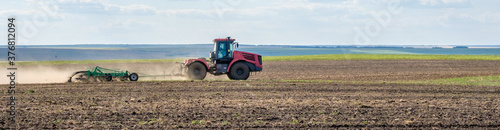 A red farm tractor in a cloud of dust cultivates the soil in the field with a cultivator after harvest. Summer sunny day. Fertile land. Modern agricultural machinery. Copy space. Banner.