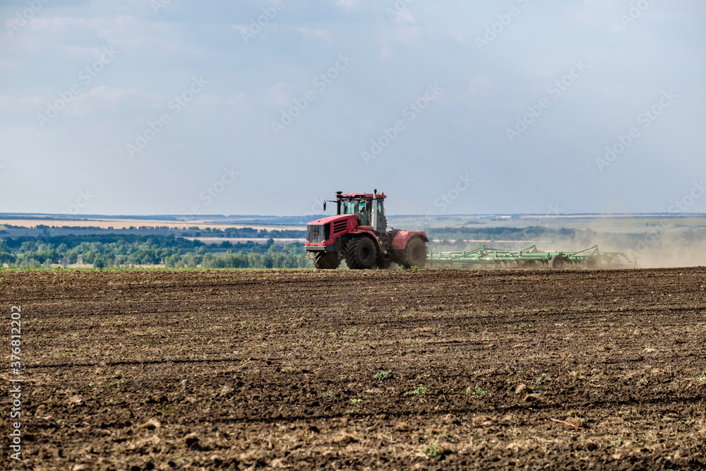 A red farm tractor in a cloud of dust cultivates the soil in the field with a cultivator after harvest. Summer sunny day. Fertile land. Modern agricultural machinery. Copy space. High quality photo A red farm tractor in a cloud of dust cultivates the soil in the field with a cultivator after harvest. Summer sunny day. Fertile land. Modern agricultural machinery. Copy space. High quality photo