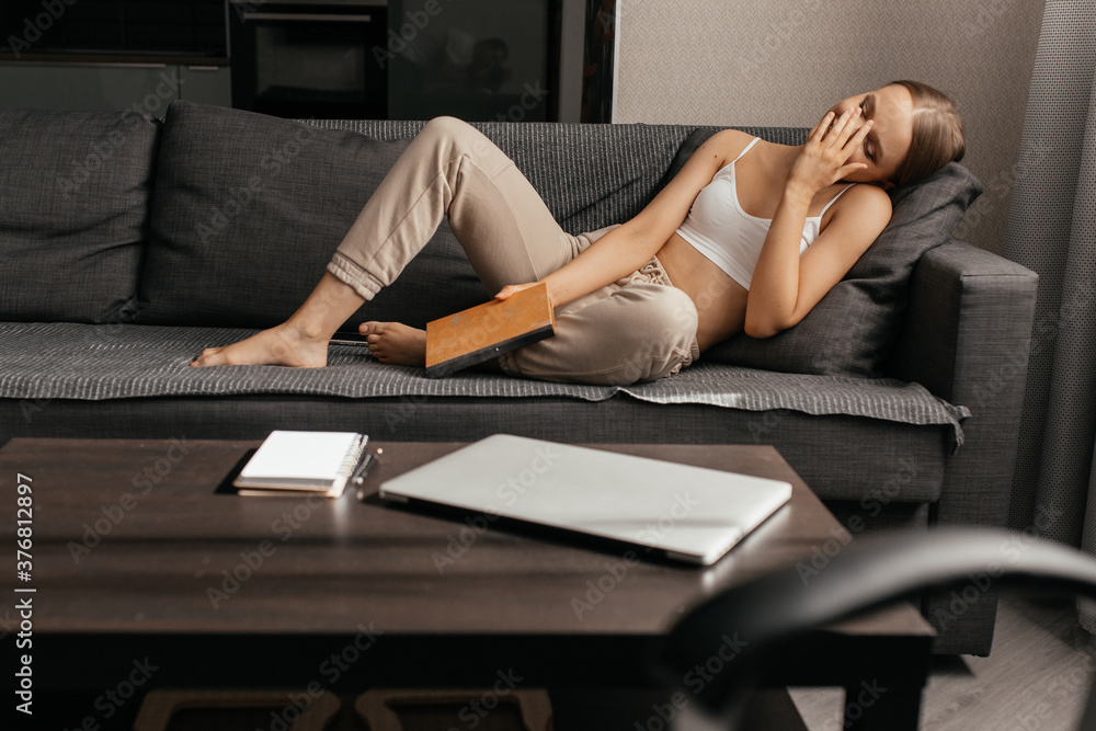 A beautiful woman in pants and a top is lying on the couch, reading a book and covering her face with her hand in surprise. Leisure at home. Development and self-study. A beautiful woman in pants and a top is lying on the couch, reading a book and covering her face with her hand in surprise. Leisure at home. Development and self-study.
