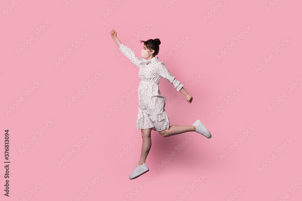 Cute brunette lady jumping on a pink studio wall gesturing like a superwoman and wear a medical mask on face Cute brunette lady jumping on a pink studio wall gesturing like a superwoman and wear a medical mask on face
