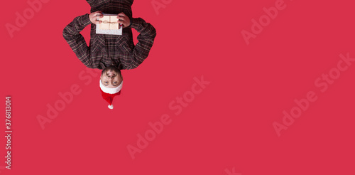 Upside down photo of a caucasian bearded man with a new year hat and present posing on a red studio wall with free space photo