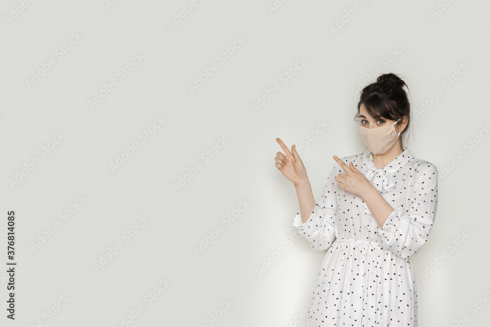 Charming brunette woman in a summer dress and medical mask on face is pointing to the white studio wall near her Charming brunette woman in a summer dress and medical mask on face is pointing to the white studio wall near her