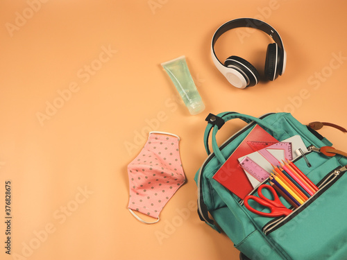 backpack with school supplies ,headphones, pink polka dot fabric masks and sanitizer gel on orange background.COVID-19 prevention while going back to school and new normal concept