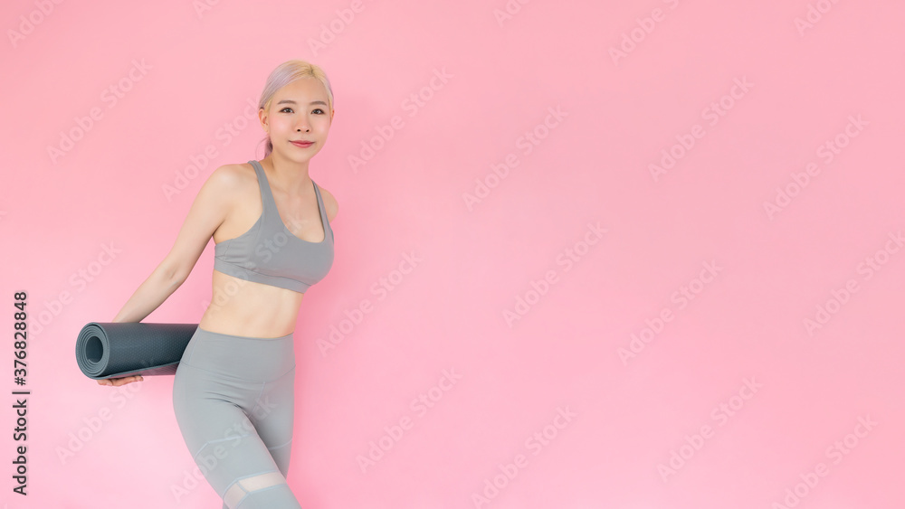 An image of a young asian woman wearing fashionable sportswear and holding a yoga mat. Female sports trainer smiling poses on a pink background. Healthy lifestyle and fitness center concept. An image of a young asian woman wearing fashionable sportswear and holding a yoga mat. Female sports trainer smiling poses on a pink background. Healthy lifestyle and fitness center concept.