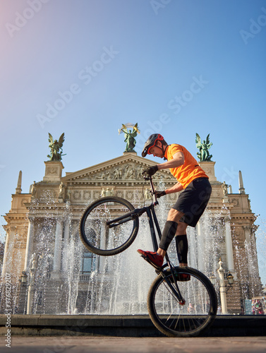 Extreme sportsman performing dangerous ride on mountain bike back wheel in front of fountain and opera house, side view, copy space