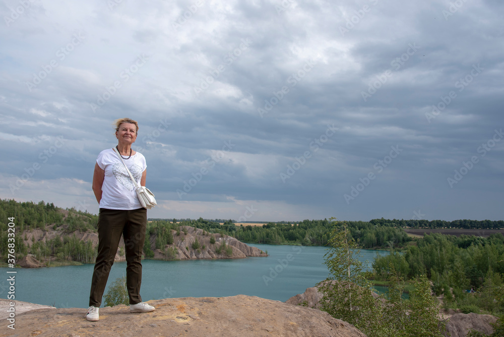 A 65-year-old woman stands on a hill against a blue lake.