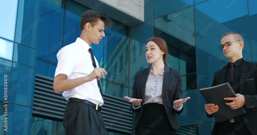Close up three successful business people stand outdoors near blue glass wall, negotiate, debate, argue. Young confident woman explains to two men knowledge information, actively moves waving hands Close up three successful business people stand outdoors near blue glass wall, negotiate, debate, argue. Young confident woman explains to two men knowledge information, actively moves waving hands