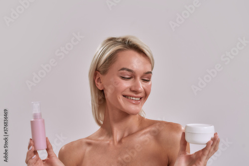 Portrait of smiling young woman with perfect skin holding jar and bottle of different cosmetic skincare products and choosing what to apply while posing isolated over grey background