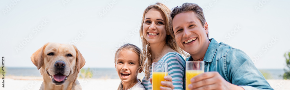 Horizontal crop of family looking at camera while holding glasses of orange juice near golden retriever on beach