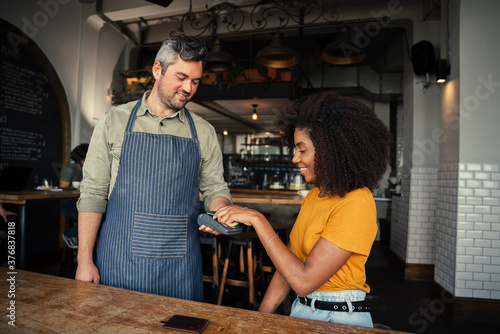 Foto Beautiful ethnic woman tapping card making payment for coffee to handsome waiter in trendy cafe