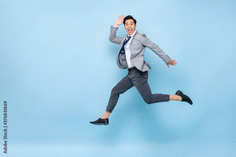 Full length fun portrait of happy energetic young Asian businessman jumping in mid-air isolated on studio blue background with copy space Full length fun portrait of happy energetic young Asian businessman jumping in mid-air isolated on studio blue background with copy space