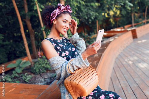 Attractive black girl in trendy hipster cardigan and headband sitting in the park and making self portrait by mobile phone .
