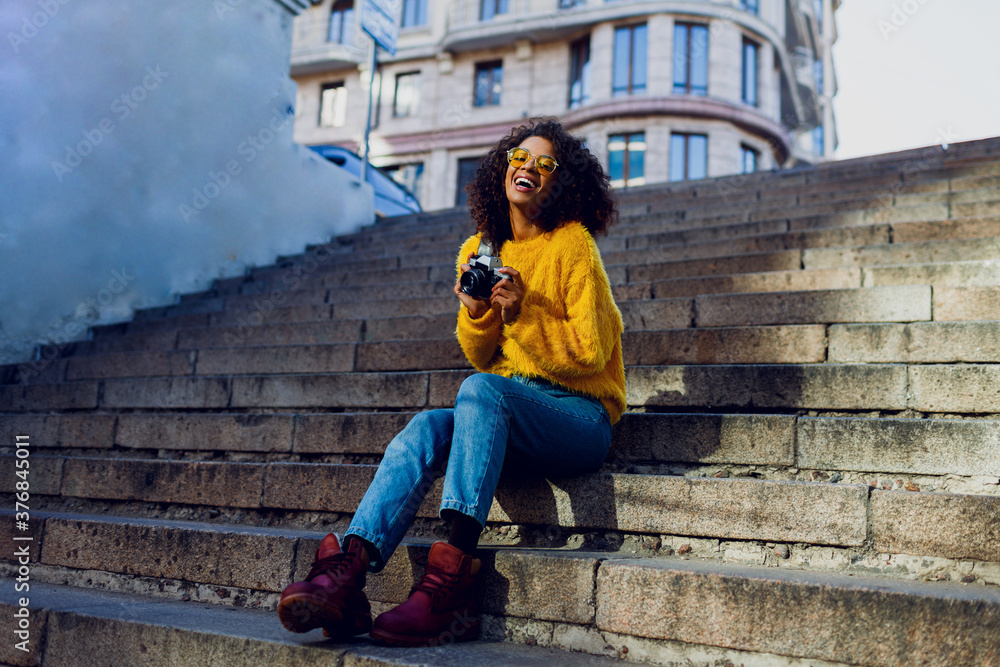 Fototapeta premium Stylish inspired American female student sitting on stairs and looking forward. . Holding retro film camera. Urban background. Autumn trends.