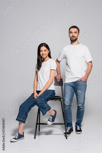 brunette asian woman in white t-shirt and jeans sitting on chair near stylish boyfriend standing with hand in pocket on grey