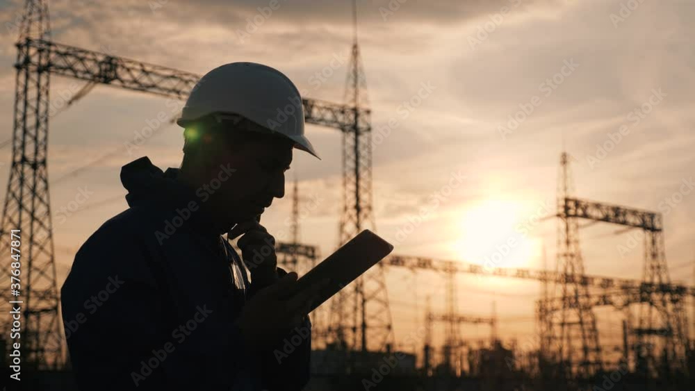 An electrical engineer wearing a helmet works with a tablet near high voltage electrical lines. An electrician works with the documents of an electrical substation. Energy business concept. An electrical engineer wearing a helmet works with a tablet near high voltage electrical lines. An electrician works with the documents of an electrical substation. Energy business concept.