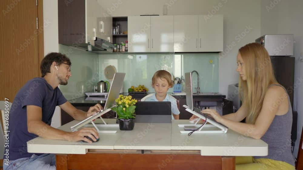 A young woman and man are working in their living room with the kitchen in the background. Their son is studying online using a tablet. They both have to work remotely due to the Covid-19 restrictions A young woman and man are working in their living room with the kitchen in the background. Their son is studying online using a tablet. They both have to work remotely due to the Covid-19 restrictions