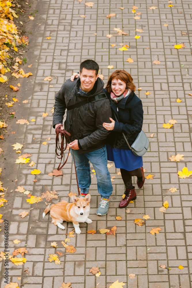 Happy couple playing with Corgi dog in sunny autumn park. Man and woman having fun outdoors with pet.