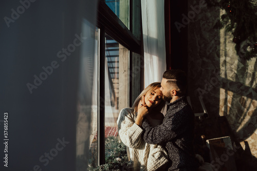 Romantic happy young couple relax at home in sunny day. They standing near big window at home.