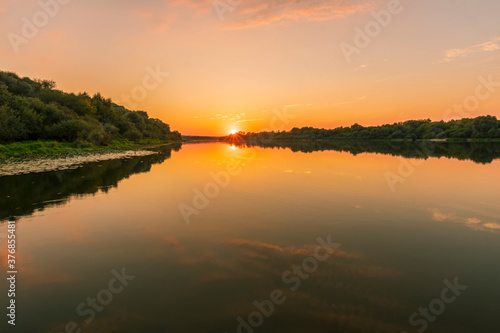 Fototapeta Naklejka Na Ścianę i Meble -  Scenic view at beautiful summer river sunset with reflection on water with trees , golden sun rays, calm water ,deep blue cloudy sky and glow on a background, spring evening landscape