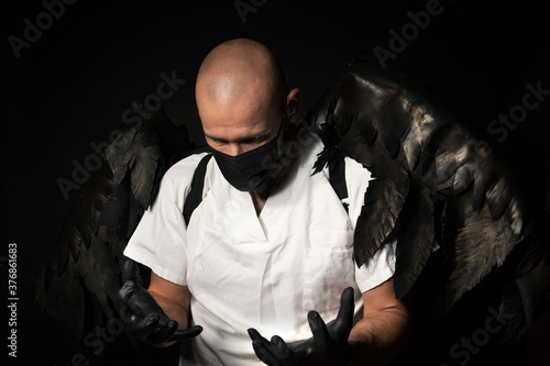 Doctor in white coat, mask, gloves and black wings, looking at hands; black wings photo