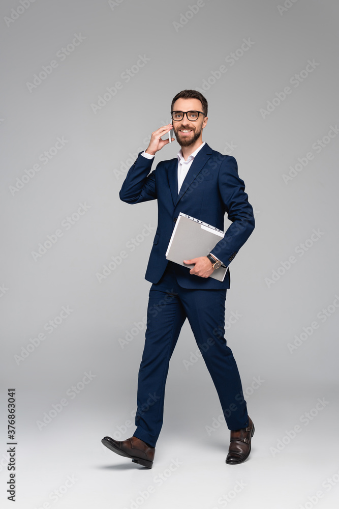 businessman in blue suit talking on smartphone and holding folder while walking on grey businessman in blue suit talking on smartphone and holding folder while walking on grey
