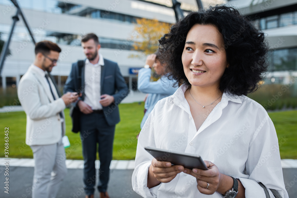 Fototapeta premium Portrait of smiling asian businesswoman with tablet standing in front of modern office buildings with colleagues behind her.