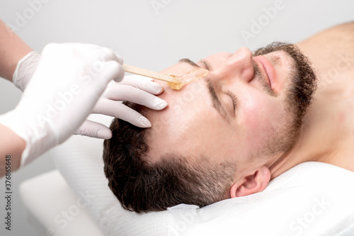 Fototapeta Naklejka Na Ścianę i Meble -  Beautician applies wax between male eyebrows before the procedure of waxing in the beauty salon