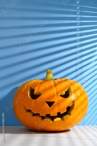 Halloween holiday background. Spooky Halloween pumpkin on a pastel blue background, illuminated by sunlight through the jalousie. Shadows. Copy space, vertical orientation. © Ramil Gibadullin