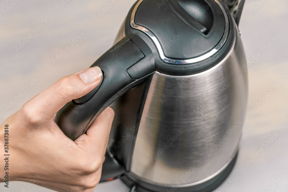 Obraz premium woman's hand holds the black handle of a gray electric kettle and presses the power button close-up, top view