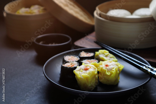 shrimp dumplings are in the black round dish with the seaweed pork rolls on the black wooden table togerther with pork and egg creamy buns in dim sum basket during lunch in Chinese Contan restaurant photo